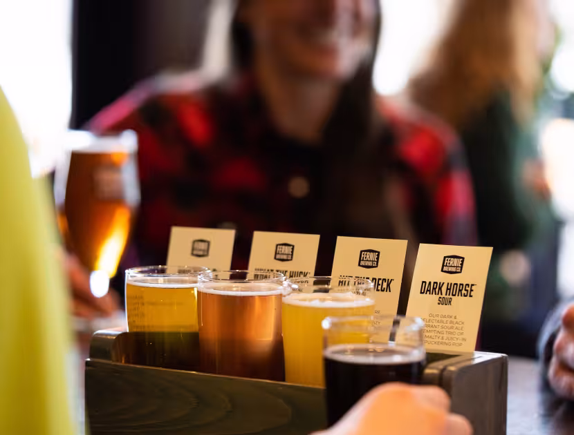 Four glasses of beer on a wooden flight paddle with blurry people in background.