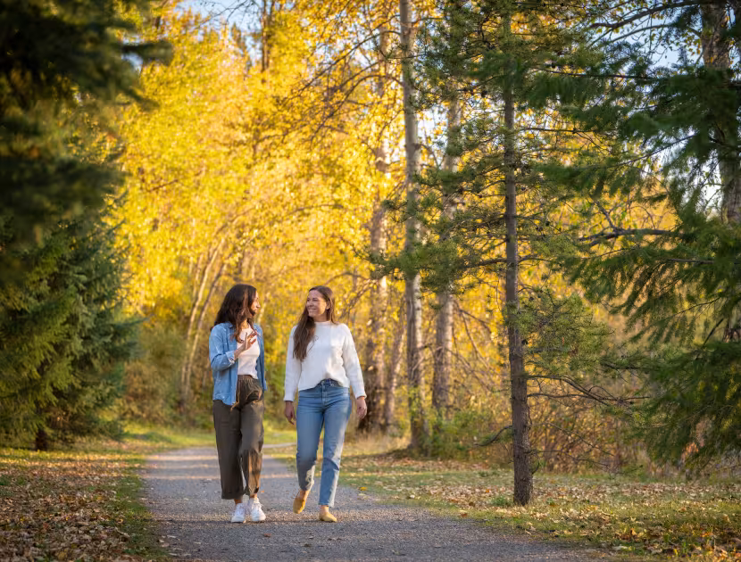 Two women walking on a path in a park with yellow and green trees.