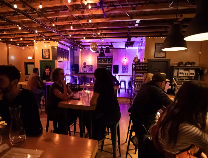Dimly lit bar interior with people seated at tables, purple lights in background.