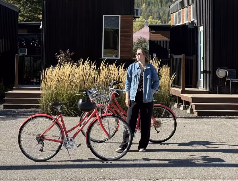 Woman standing beside a red bicycle with modern homes and a mountain in the background.