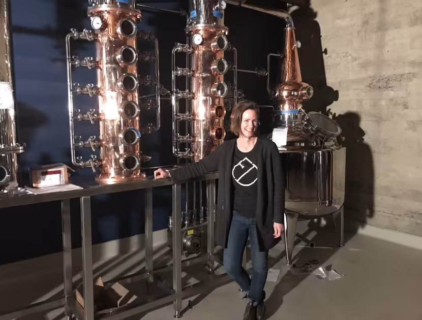 Woman standing beside copper distillation equipment.