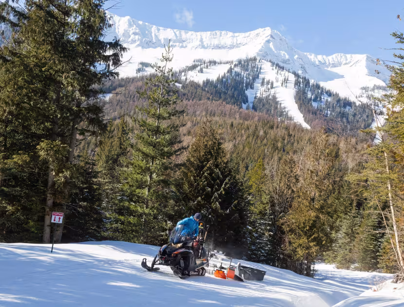 Groomer on a snowmobile grooming a trail with snowy mountains and trees in the background.