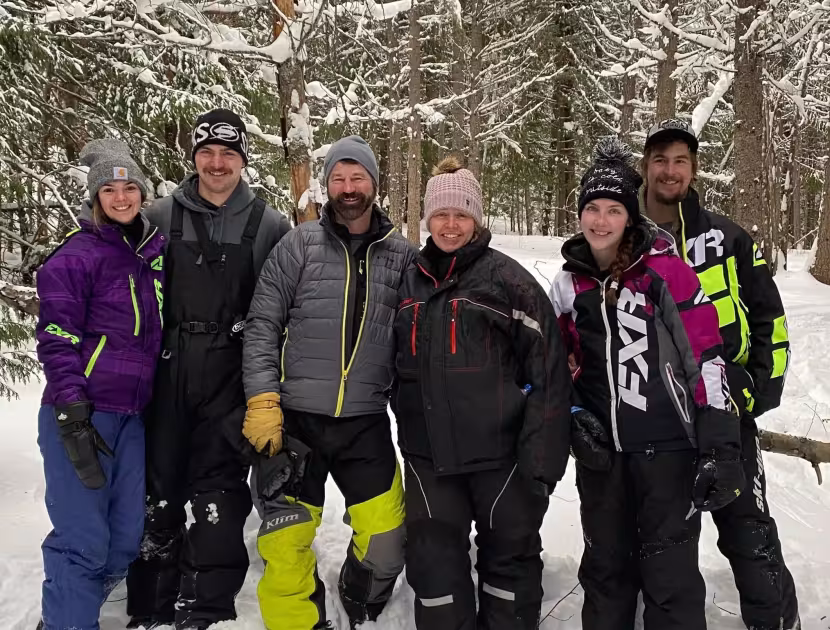 Six people in winter gear standing together in snowy woods.