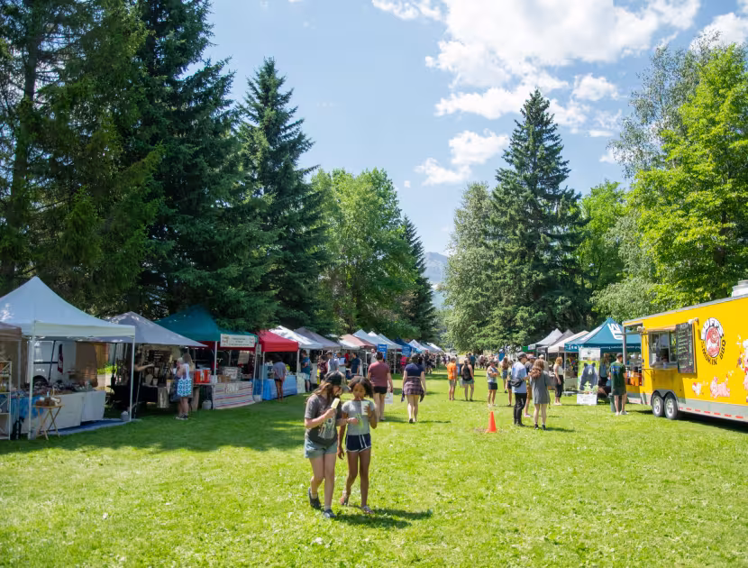 Outdoor farmers market with people, tents, and a yellow food truck on grassy field.
