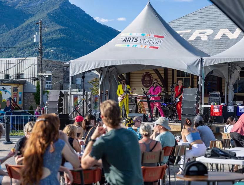 People sitting at tables watching a band perform on an outdoor stage under a tent, with mountains in the background.