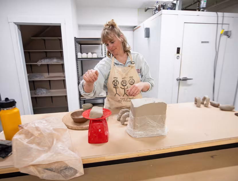 Woman in apron working with clay on a wooden table in a studio.