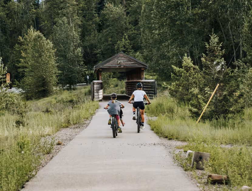 A family of three riding on a path through a grassy area with trees and a wooden bridge in the background.