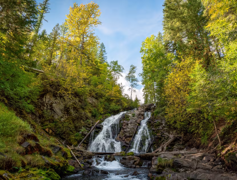 Waterfall cascading down rocky slope, surrounded by lush greenery and trees; blue sky with clouds in background.