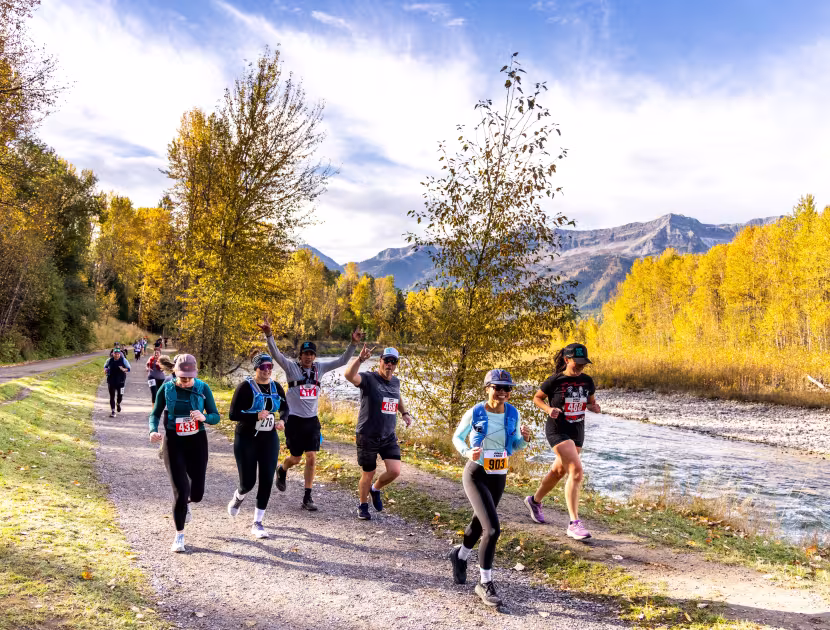 Runners on a path by a river, surrounded by autumn trees and mountains.