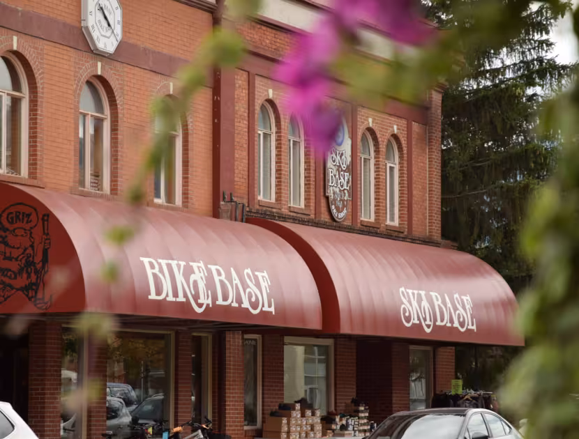 Red brick building with clock and awning reading "Ski Base" and "Bike Base", cars parked outside.