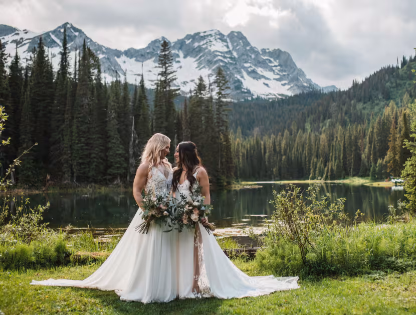 Two women in white wedding dresses embracing, holding bouquet, in front of serene lake and snow-capped mountains.