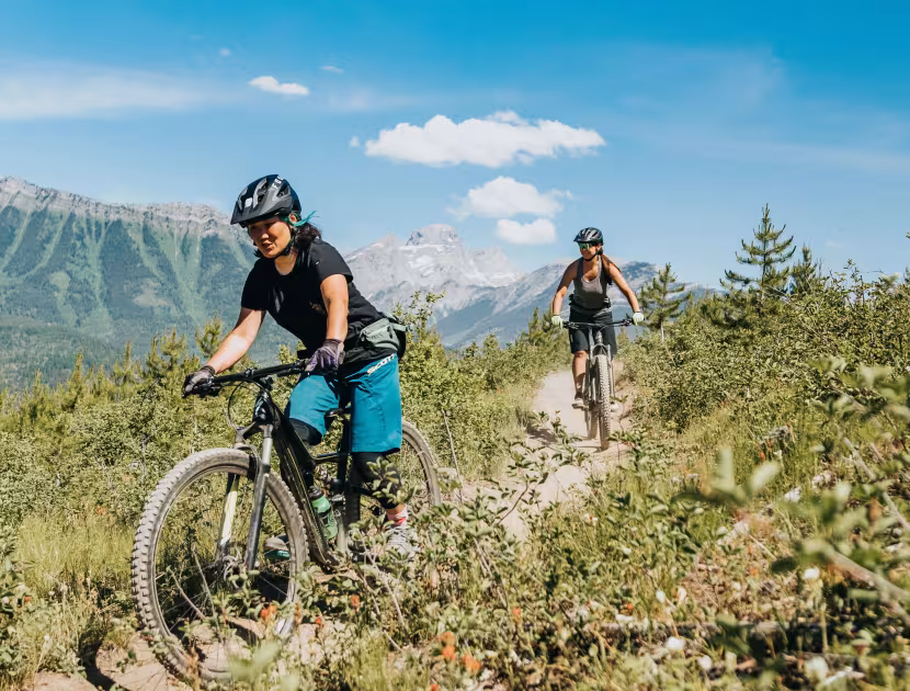 Two cyclists ride mountain bikes on a trail through grassy terrain with mountains and blue sky in the background.