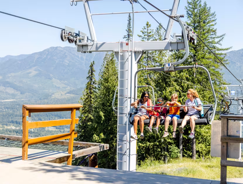 Two adults and two children riding a ski lift on a sunny summer day with mountains and trees in the background.