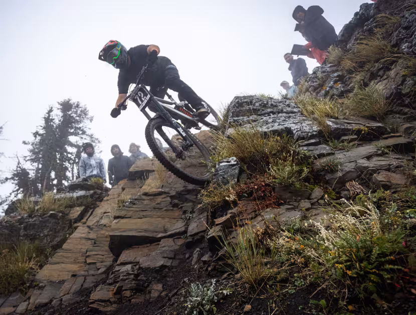 Mountain biker navigating rocky trail with spectators; overcast sky.