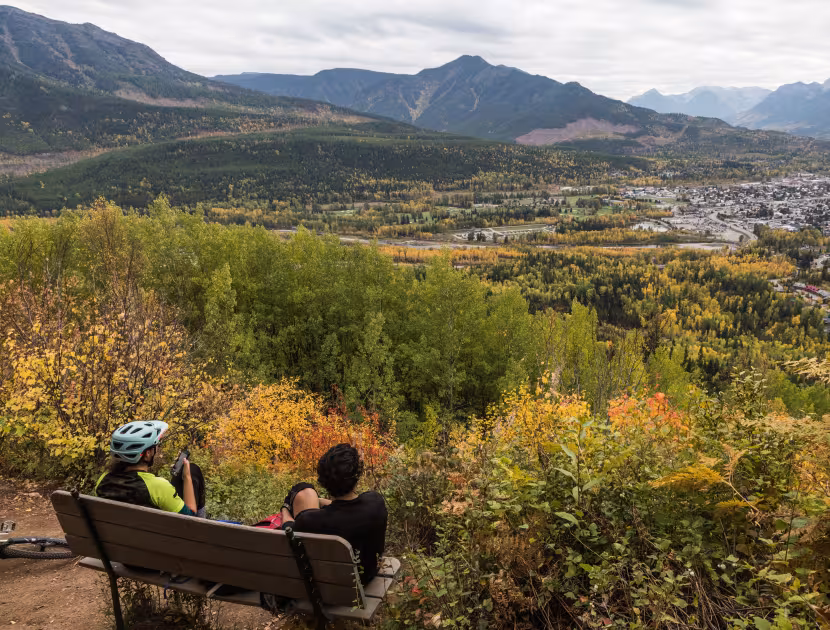 Two mountain bikers sitting on a bench, overlooking a valley with mountains and trees changing colors.