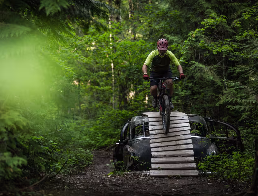 Man on a mountain bike riding over a wooden bridge over a vintage black car in forest.