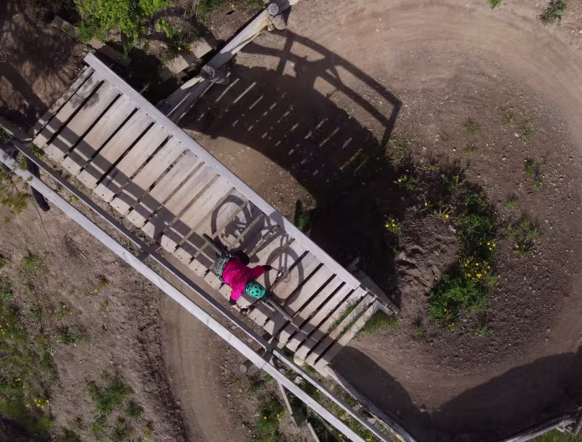 Aerial view of a wooden bridge leading to a fenced dirt area with sparse greenery; a person in pink walks on the bridge.
