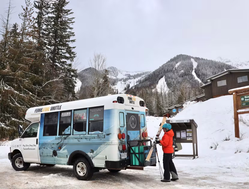 Person in orange jacket opening back of blue and white ski shuttle minibus on snowy road with mountains and trees in background.