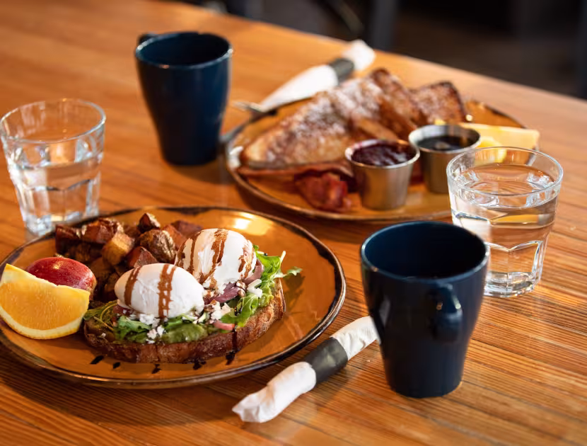 Two plates of food with toast, potatoes, and orange slices; two glasses of water; two black mugs on wooden table.