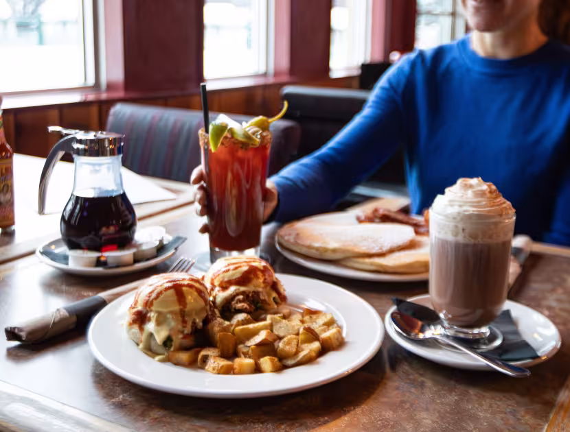 Breakfast table with pancakes, eggs, coffee, and syrup; woman in blue sweater sits in background.