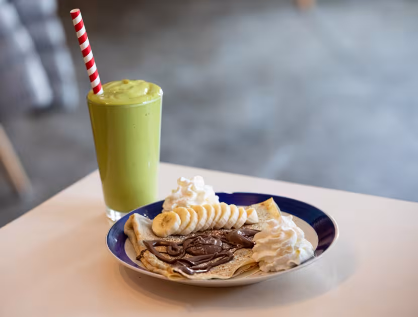 A plate of crepes with chocolate sauce and a green smoothie on a table.