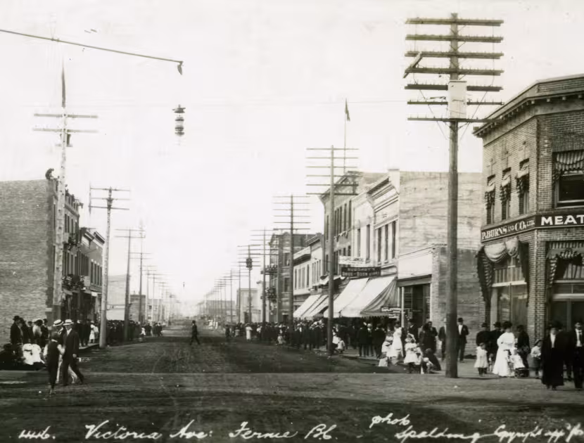 Historic black and white photograph of a street scene in Fernie with people walking, storefronts, and power lines.