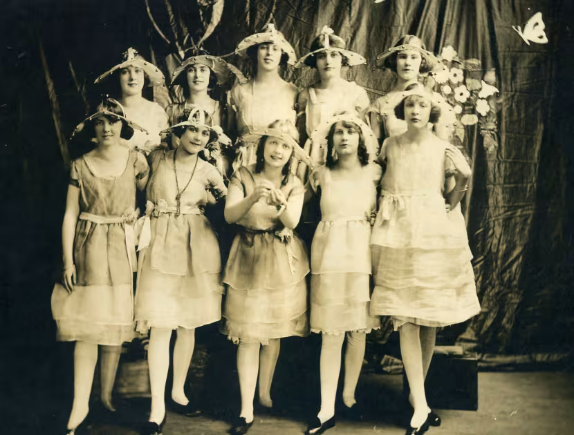 Historic black and white photograph of nine young women in dresses and hats, posed in front of a draped backdrop.