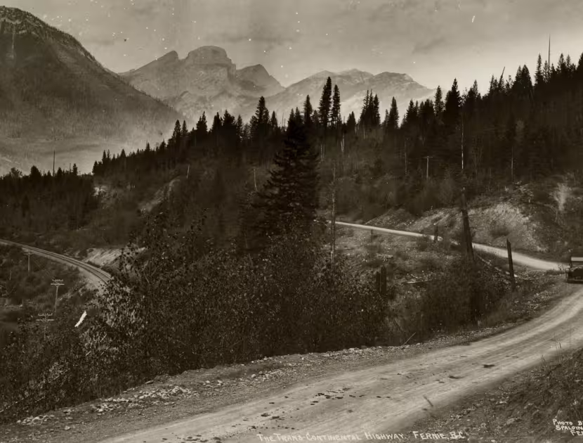 Historic black and white photograph of the Transcontinental Highway in Fernie, a winding road through a forested mountainous area with a car driving away.