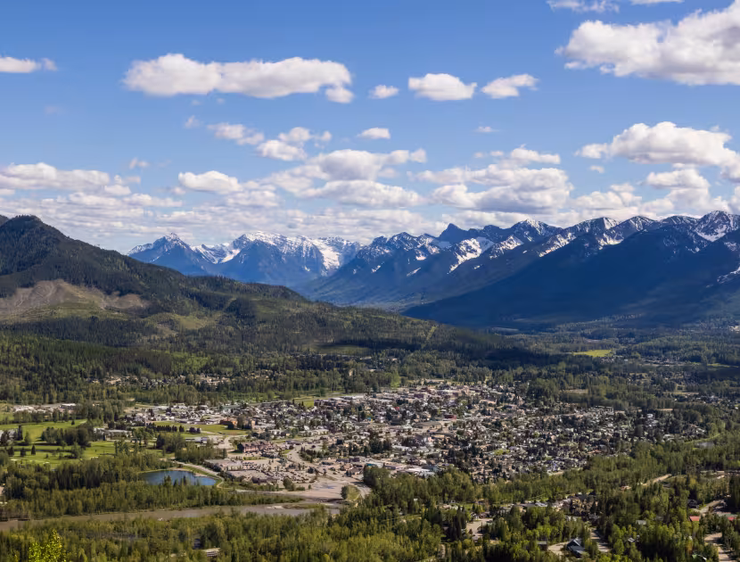 Panoramic view from the Swine Flu bench of a Fernie surrounded by green mountains with snow-capped peaks under a blue sky with white clouds.