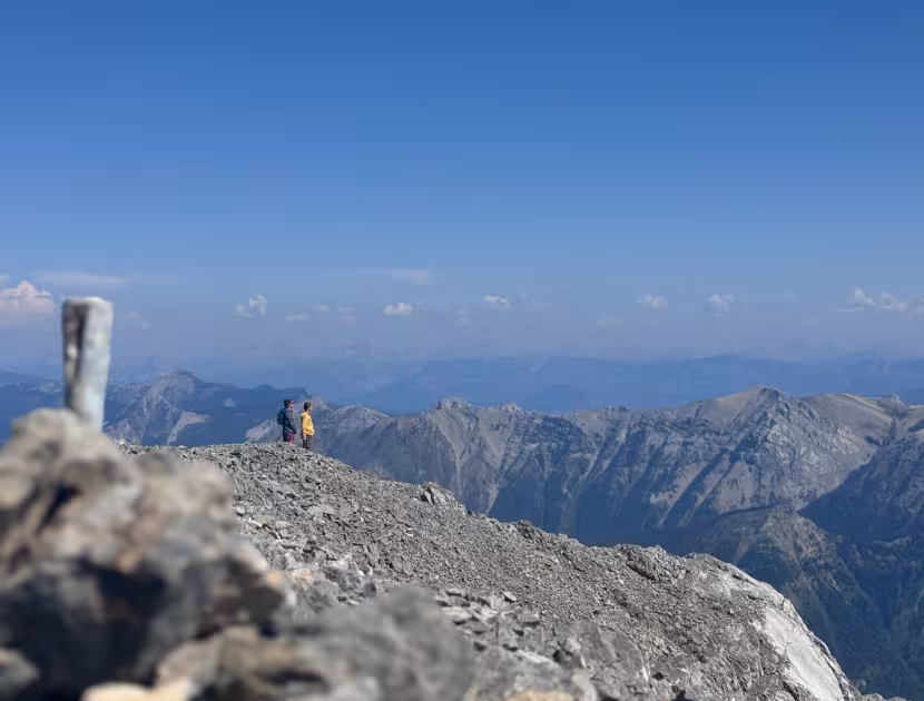 Two people standing on the peak of the Three Sisters Mountain in Fernie with vast mountain range and blue sky.