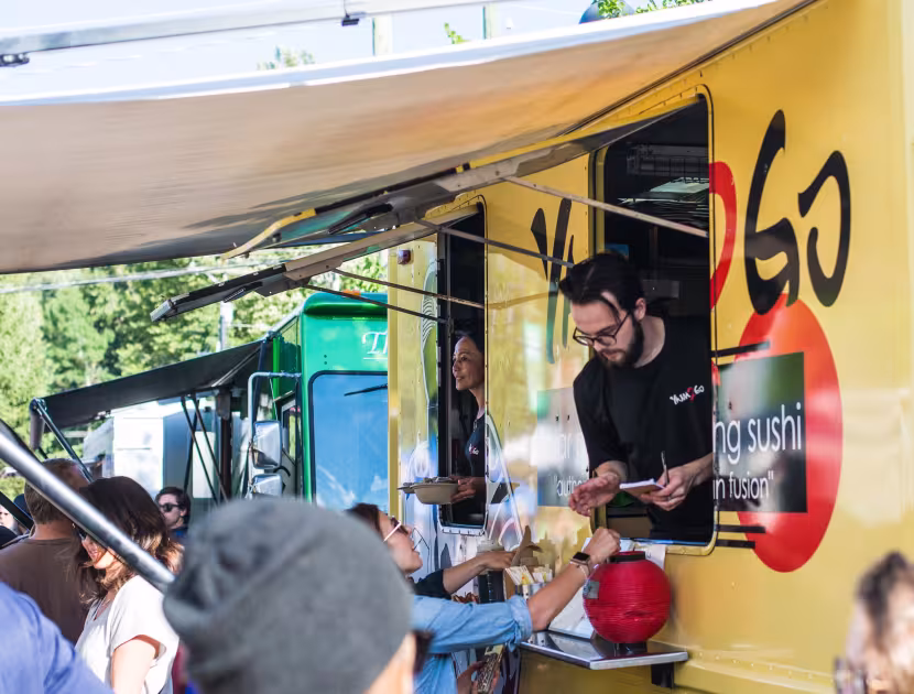 Man serving food from a yellow food truck window; crowd gathered.