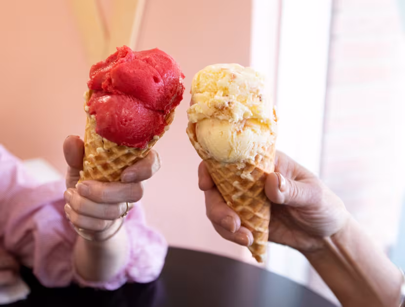 Two hands holding ice cream cones, one with red ice cream and one with yellow.