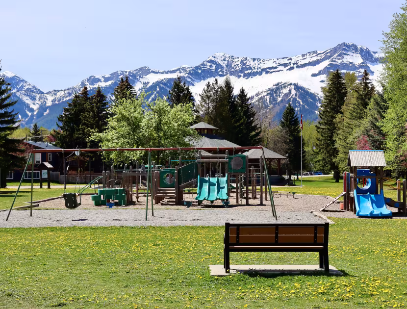 Rotary Park in Fernie with a playground with swings and slides on a sandy patch surrounded by grass, trees, and a mountain range in the background.