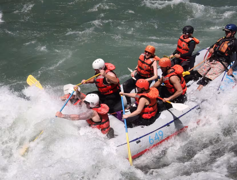 People in red life jackets and helmets navigate a white raft through turbulent water, holding yellow and blue paddles.
