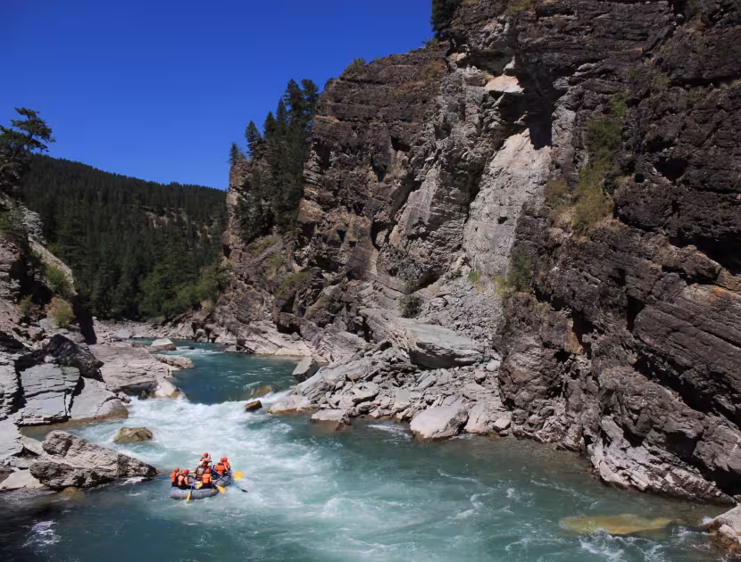 Rafters on a river flowing through a rocky gorge with trees on the left, under a clear blue sky.