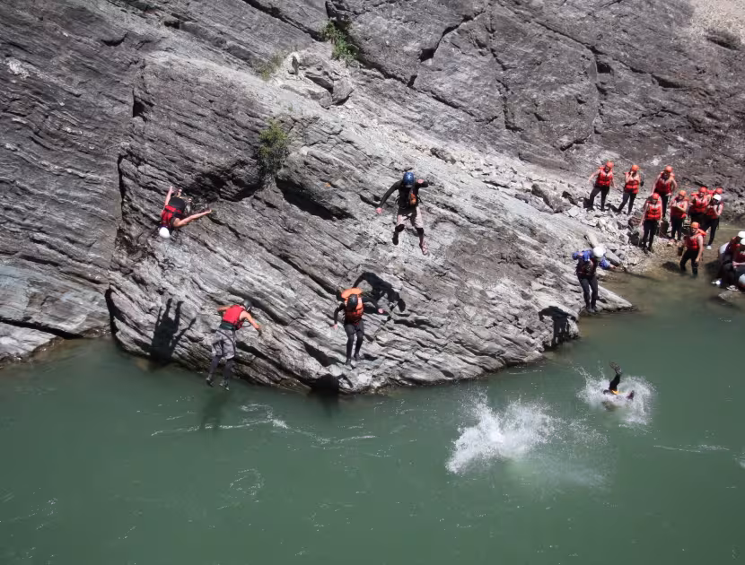People in orange life jackets jumping off a cliff into green water.