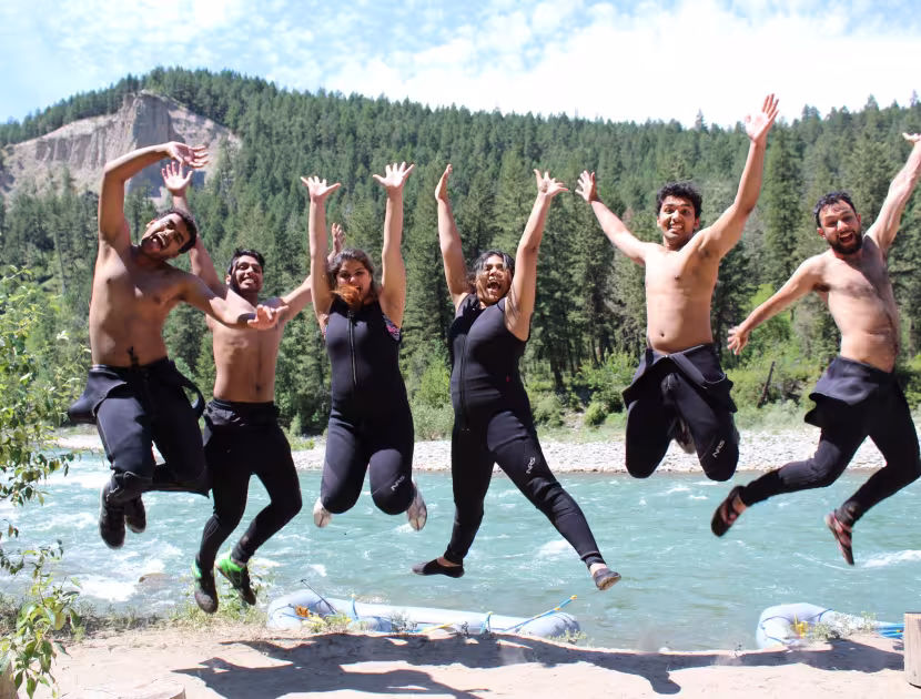 Six people jumping in unison by a river, wearing black wetsuits, with a forested hill in the background.