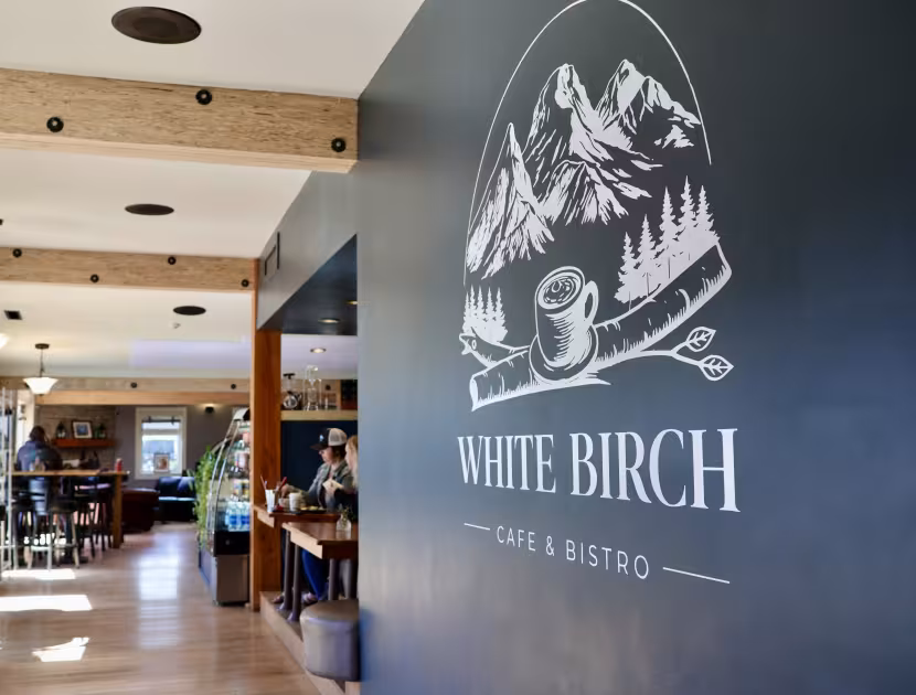 A logo on a navy blue wall featuring a white illustration of mountains and trees with a mug and utensils, reading "WHITE BIRCH CAFE &amp; BISTRO".
