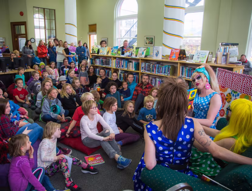 Children sitting in a circle on floor of a library, listening to two drag queens.