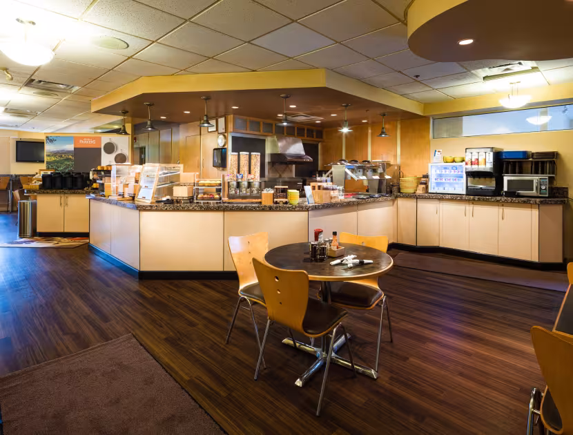 Cafeteria with a table and chairs in the foreground; kitchen area with counter and microwaves in background.