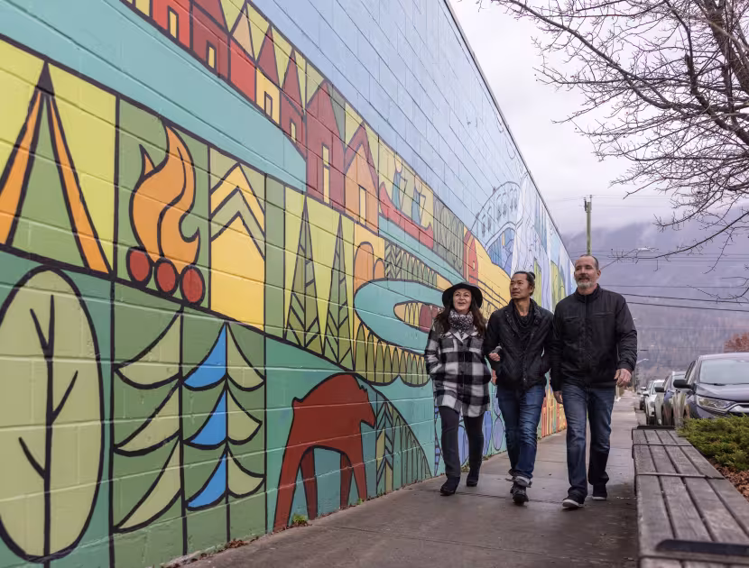Three people walking past a colorful mural on a sidewalk.