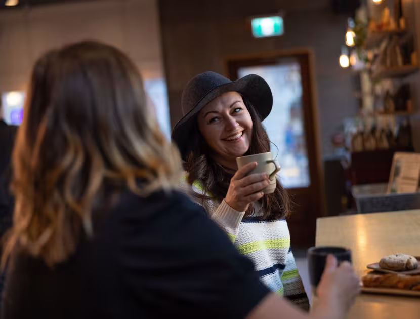 Two women sitting in a coffee shop; one smiling and holding a coffee mug.
