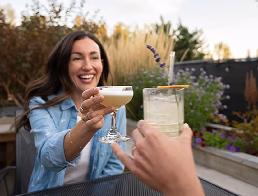 Woman toasting a drink with someone outdoors, smiling and sitting in a garden.