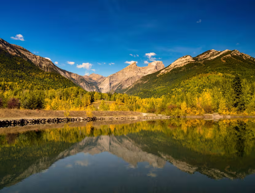 Three Sisters mountain range reflected in still Maiden lake water, surrounded by green and yellow trees under a blue sky.
