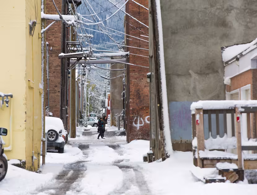 Snow-covered urban alley with brick buildings, utility wires, and a wooden porch.