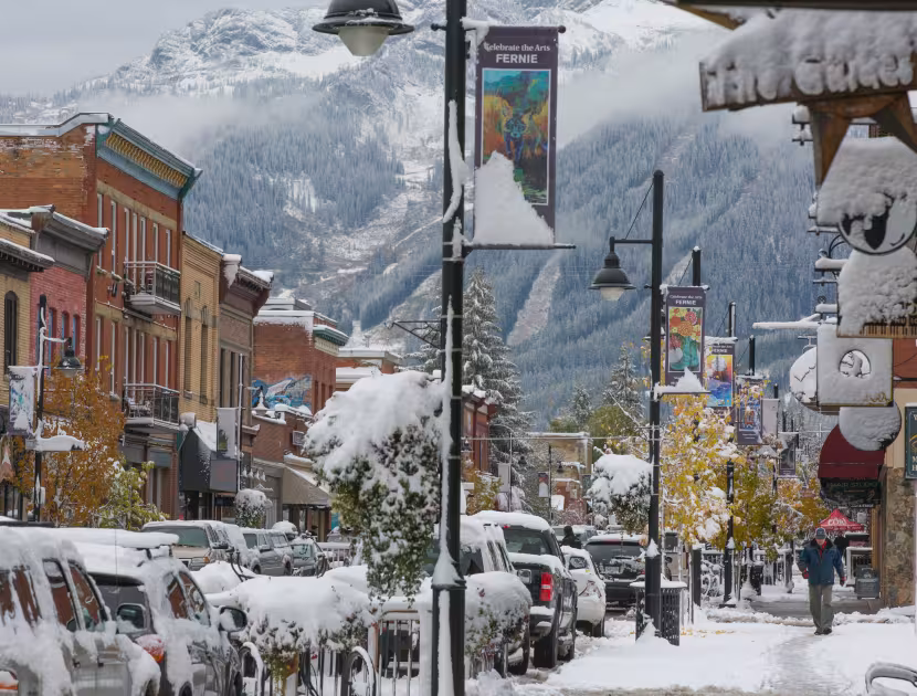 Snow-covered mountain town street with parked cars and colorful buildings.