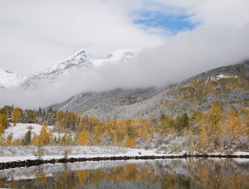 Snowy mountains and autumn trees reflected in a calm lake beneath low clouds.