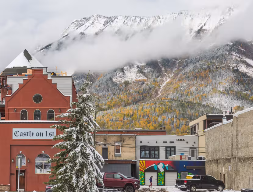 Snowy mountain town with brick buildings, evergreen trees, and low clouds.