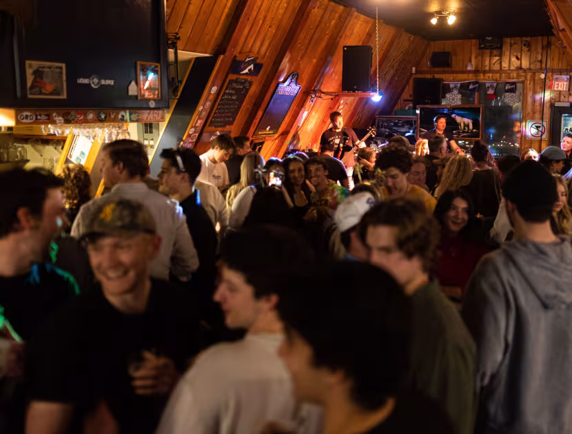Crowded bar with people socializing under warm wooden beams and neon lights.
