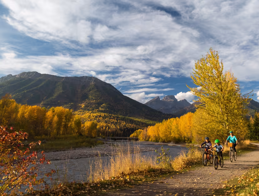 Cyclists ride along a lakeside trail beneath autumn mountains and cloudy skies.